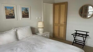 Um quarto com cama, mesa de cabeceira, abajur, porta de madeira, espelho fixado na parede e duas fotos emolduradas.