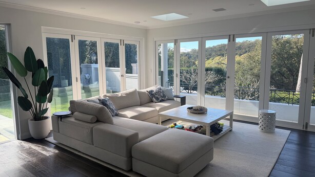 Uma sala de estar moderna com um sofá seccional, uma mesa de centro e uma janela grande com vista para um jardim e árvores.