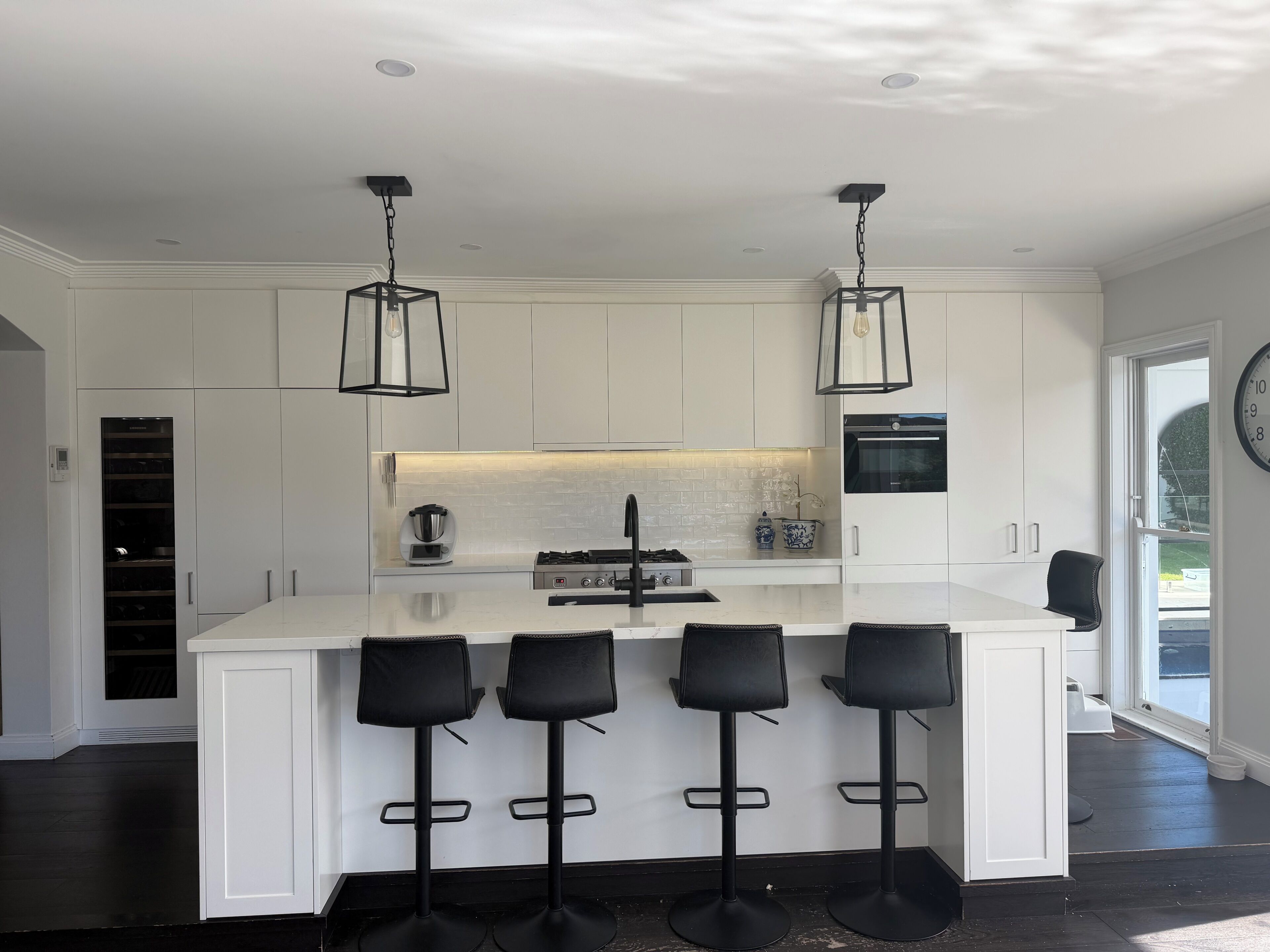 A modern kitchen with white cabinetry, a central island with bar stools, pendant lighting, and a visible clock on the wall.