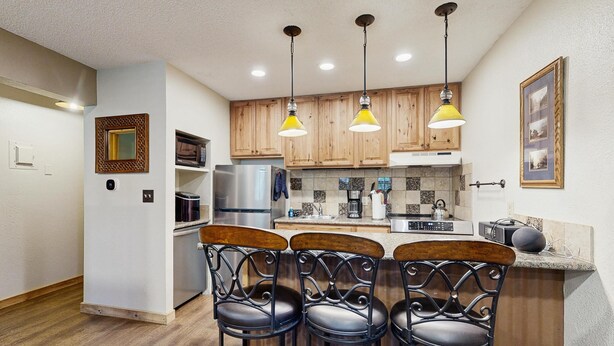 A kitchen with wooden cabinets, a stainless steel refrigerator, a coffee maker, and a bar with three stools. - Conveniently located condo w/ fast wifi, great location & shared pool & hot tub (Winter Park)