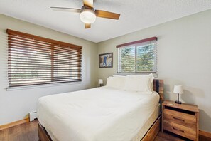 Une chambre à coucher comprenant un lit, une table de chevet, une lampe, un ventilateur de plafond et deux fenêtres équipées de stores.