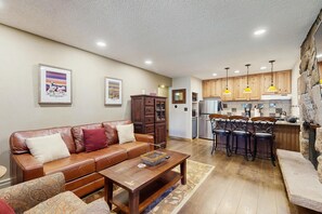 Un salon avec un canapé en cuir marron, une table basse en bois et un coin cuisine doté de meubles en bois et d’un réfrigérateur en acier inoxydable.