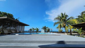 A resort pool area with a clear view of the ocean, surrounded by palm trees and a wooden fence.