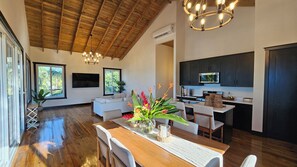 A spacious living area with a wooden ceiling, a dining table with chairs, a kitchen with dark cabinets, and a large window. - Casa Buenas Vibras 5-bedroom Beach Front house in Rio Maria (Rio Maria)