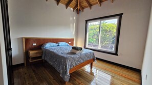 A bedroom with a wooden headboard, a bed with a floral bedspread, a bench, and a window with a view of trees. - Casa Buenas Vibras 5-bedroom Beach Front house in Rio Maria (Rio Maria)