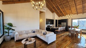 A spacious living area with a large L-shaped sofa, a round coffee table, and a dining table with chairs. There is a kitchen area with modern appliances and a large window providing a view. - Casa Buenas Vibras 5-bedroom Beach Front house in Rio Maria (Rio Maria)