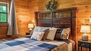 Ein Zimmer mit Holzwänden, einem Bett, zwei Nachttischlampen, einem Fenster mit Vorhang und einem an der Wand befestigten Kranz.