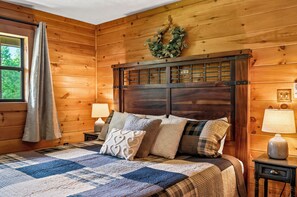 A wooden-paneled room with a bed, two bedside lamps, a window with a curtain, and a wall-mounted wreath. - New! Dollywood~Secluded~Hot Tub~King Suite~Firepit (Sevierville)