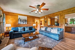 L’intérieur d’une cabane en rondins, avec deux canapés, une table basse, un ventilateur de plafond et un coin cuisine visible à travers une porte ouverte.