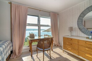 A bedroom with a large window, a wooden desk, a wicker chair, a rattan dresser, and a round mirror.
