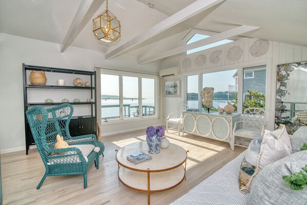 A spacious room with a large window overlooking a body of water, a round table, and a wicker chair.