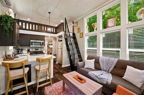 A modern kitchen with a central island, wooden bar stools, a dining area, a living room with a sofa and a coffee table, and a staircase leading to an upper level. - New! Dollywood, Views, Hot Tub, Fireplace, Easy Rd (Sevierville)