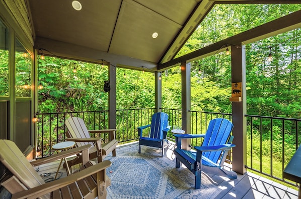 A covered outdoor seating area with Adirondack chairs and a small table, surrounded by a railing with a view of dense greenery.