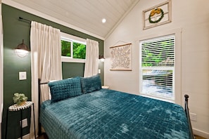 A bedroom with a bed, curtains, a wall-mounted light fixture, a window with blinds, and a decorative wreath on the wall.