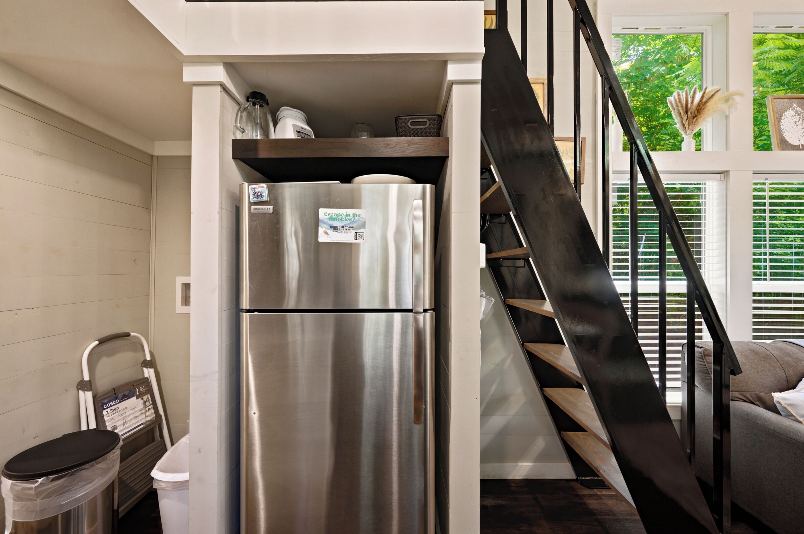 A compact kitchen with a stainless steel refrigerator, a trash bin, and a staircase leading to an upper level.