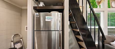 A compact kitchen with a stainless steel refrigerator, a trash bin, and a staircase leading to an upper level.