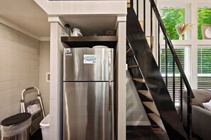 A compact kitchen with a stainless steel refrigerator, a trash bin, and a staircase leading to an upper level.