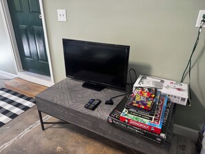 A television on a wooden stand with a remote control, a black and white checkered rug, and a stack of board games including UNO and Monopoly. - Adorable 2-bedroom condo in charming Arden near airport (Arden)