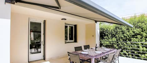 Eine Terrasse im Freien mit einem überdachten Esstisch, einer Tür zu einem Innenraum und Blick auf Grün im Hintergrund.