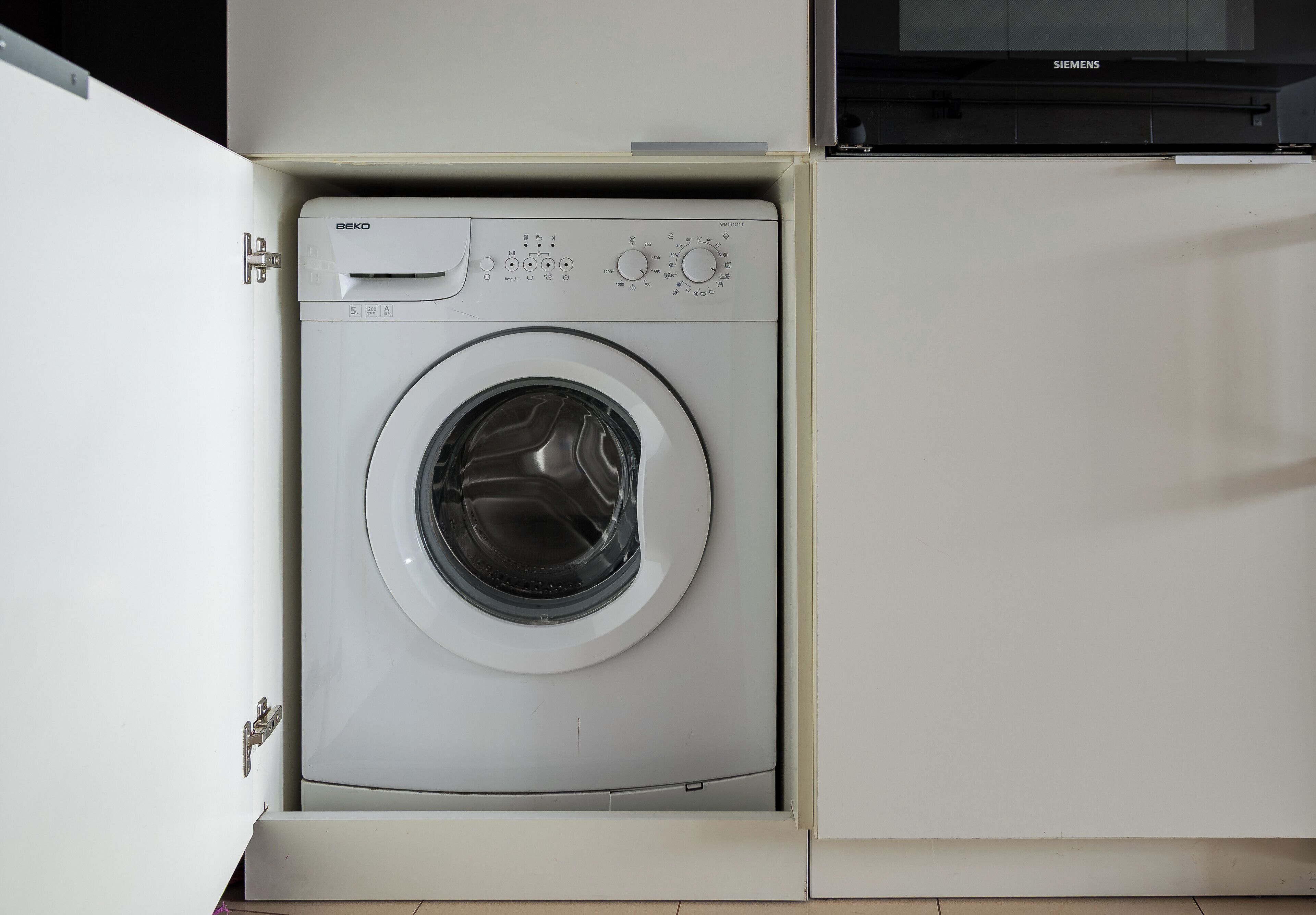 A front-loading washing machine in a white cabinet with a control panel and a Siemens brand microwave above it.