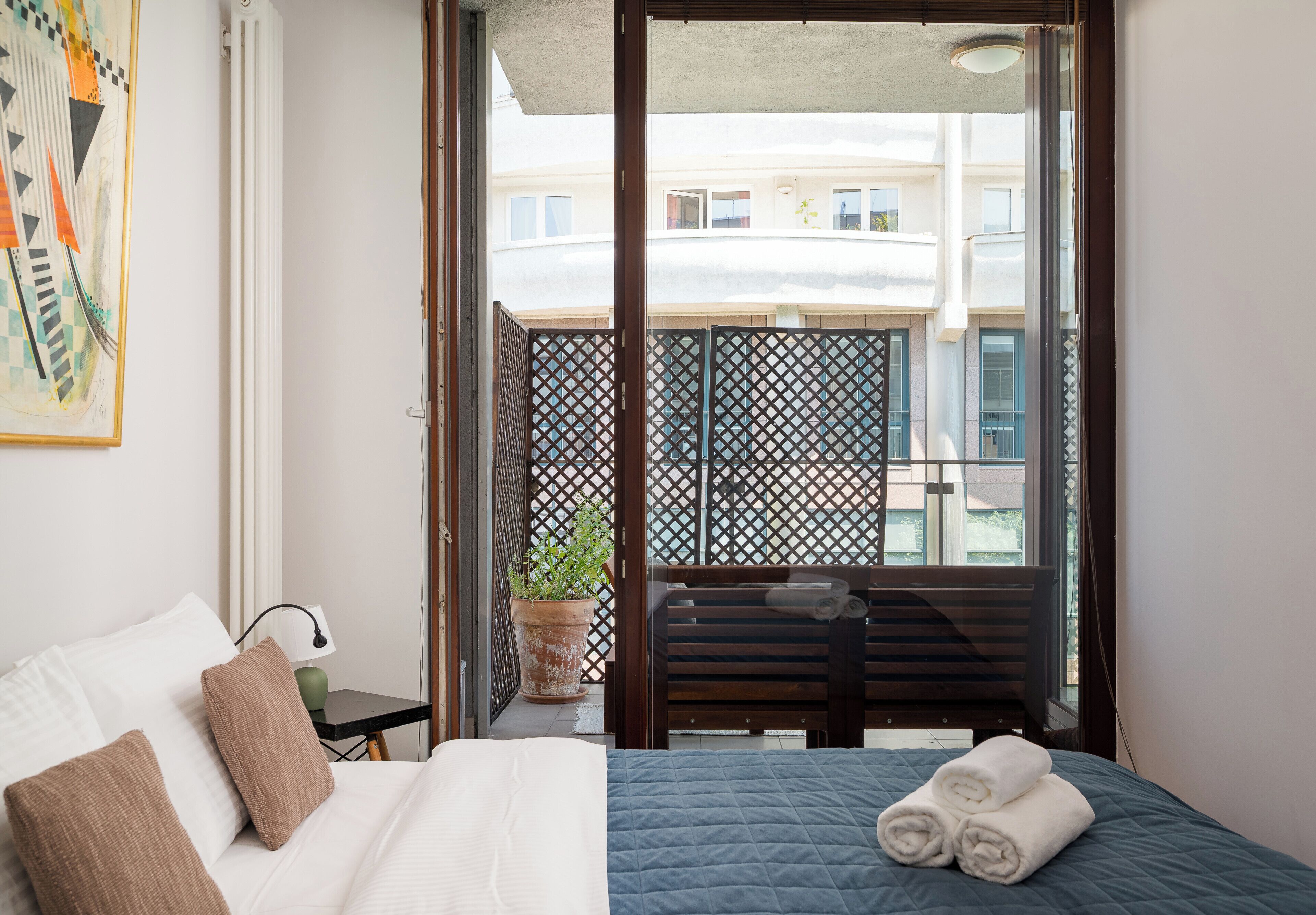Habitación de hotel con una cama, almohadas, una mesita con una lámpara, una planta en maceta y un balcón con vistas a edificios.