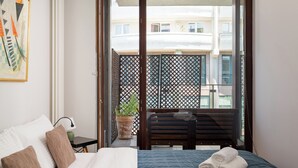 A hotel room with a bed, pillows, a small table with a lamp, a potted plant, and a balcony with a view of buildings. - Ochota Superior Apartment (Warszawa)