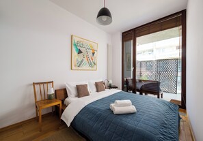 Ein Schlafzimmer mit einem Bett, einem Sessel, einem Nachttisch, einer Lampe, einem Bild und Blick ins Freie.