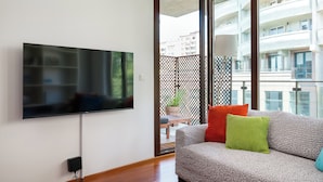 A modern living room with a flat-screen TV mounted on the wall, a sofa with cushions, and a balcony with a view of buildings. - Ochota Superior Apartment (Warszawa)