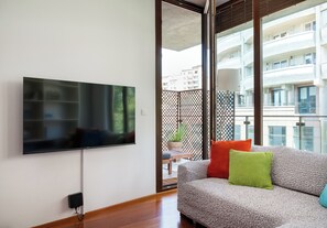 Ein modernes Wohnzimmer mit einem wandmontierten Flachbildfernseher, einer Couch mit Kissen und einem Balkon mit Blick auf Gebäude.