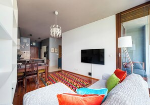 Una sala moderna con un televisor de pantalla plana, un comedor con una mesa y sillas de madera, y una alfombra colorida.