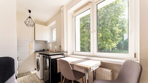 Une petite cuisine équipée d’un lave-linge, d’une table et de chaises, d’un radiateur et d’une fenêtre donnant sur des arbres.