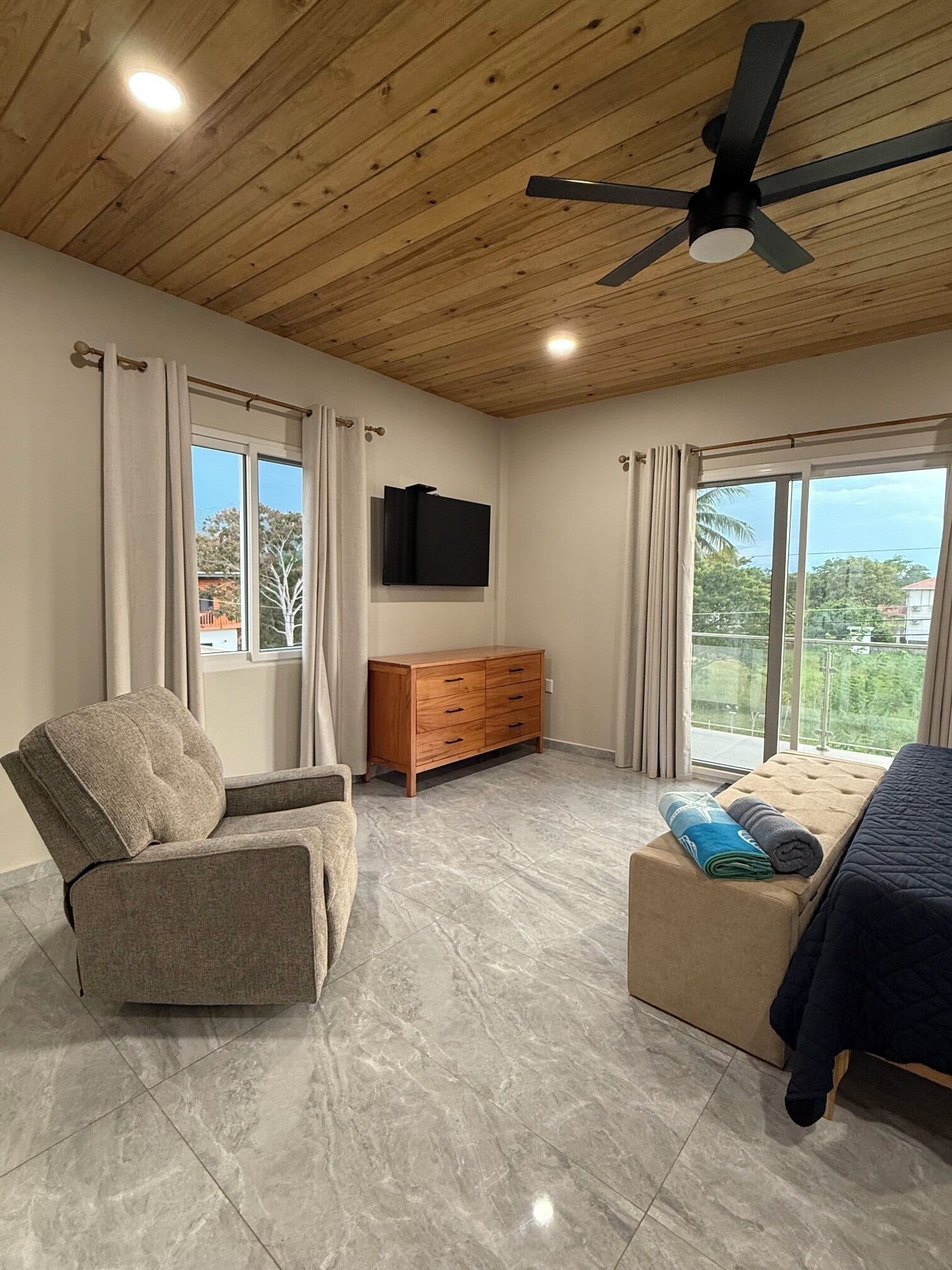 Un dormitorio moderno con ventilador de techo, televisor montado en la pared, mueble de madera, sofá, cama con manta azul y vistas por la ventana grande.