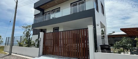 A two-story modern house with a balcony, a gated entrance, and a paved driveway.