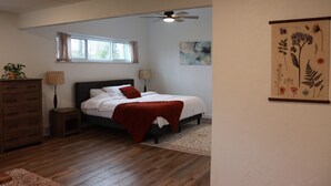 A bedroom with a bed, a nightstand, a dresser, a ceiling fan, a window with curtains, and a wall with a floral painting.