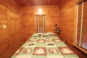 Ein Zimmer einer Holzhütte mit einem Bett, einem Nachttisch und einem Fenster mit Jalousien.