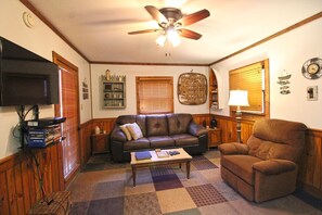 A living room with a brown leather sofa, a brown armchair, a wooden coffee table, a ceiling fan, a flat-screen TV, and a wooden cabinet. - Creekside Treehouse- Creekside Cabin (Bryson City)