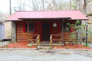 Eine Blockhütte mit rotem Metalldach, einer Holzveranda und einem kleinen Vorgarten mit Mulch und trittfesten Steinen.