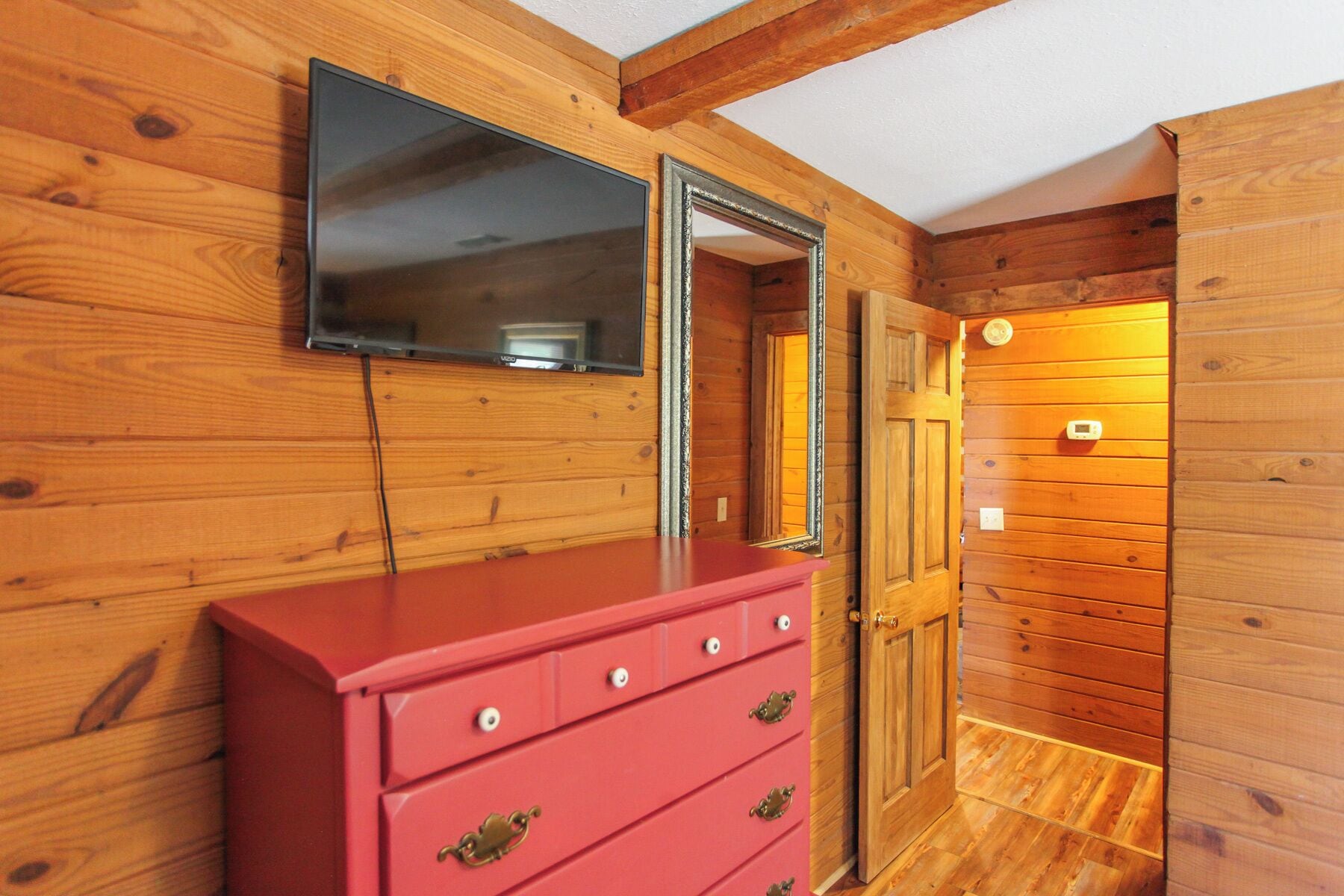 A wooden interior with a red dresser, a flat-screen TV mounted above it, and a door leading to another room.