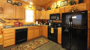 A wooden cabin-style kitchen with wooden cabinets, a black refrigerator, a microwave, a stove, and a dishwasher. - Trout Creek Cabin- Right on the Creek (Whittier)