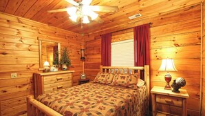 A wooden cabin bedroom with a bed, nightstands, a ceiling fan, and a window with curtains. - Trout Creek Cabin- Right on the Creek (Whittier)