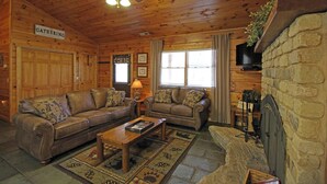 A log cabin interior with a fireplace, two sofas, a coffee table, and a ceiling fan. - Trout Creek Cabin- Right on the Creek (Whittier)