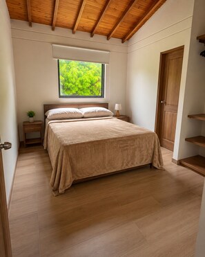 Quarto com uma cama, uma mesa de cabeceira, janela com vista para vegetação e teto de madeira.