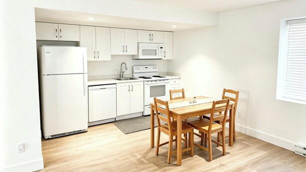 A compact kitchen with white cabinets, a refrigerator, a microwave, and a stove. There is a wooden dining table with chairs. - A bright, clean walking-out basement unit in Surrey,near White-rock. (Surrey)