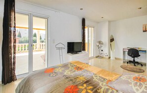 Ein Hotelzimmer mit einem Bett, einem Schreibtisch mit Stuhl, einem Fernseher, einem Ventilator und einem großen Fenster mit Blick auf Grünflächen.