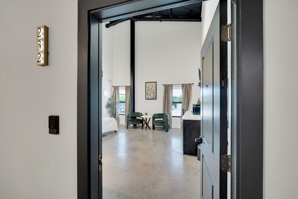 A hotel room with a bed, two chairs, a small table, a desk, a painting on the wall, and a door with a number sign. - The Lawson District (Midlothian)