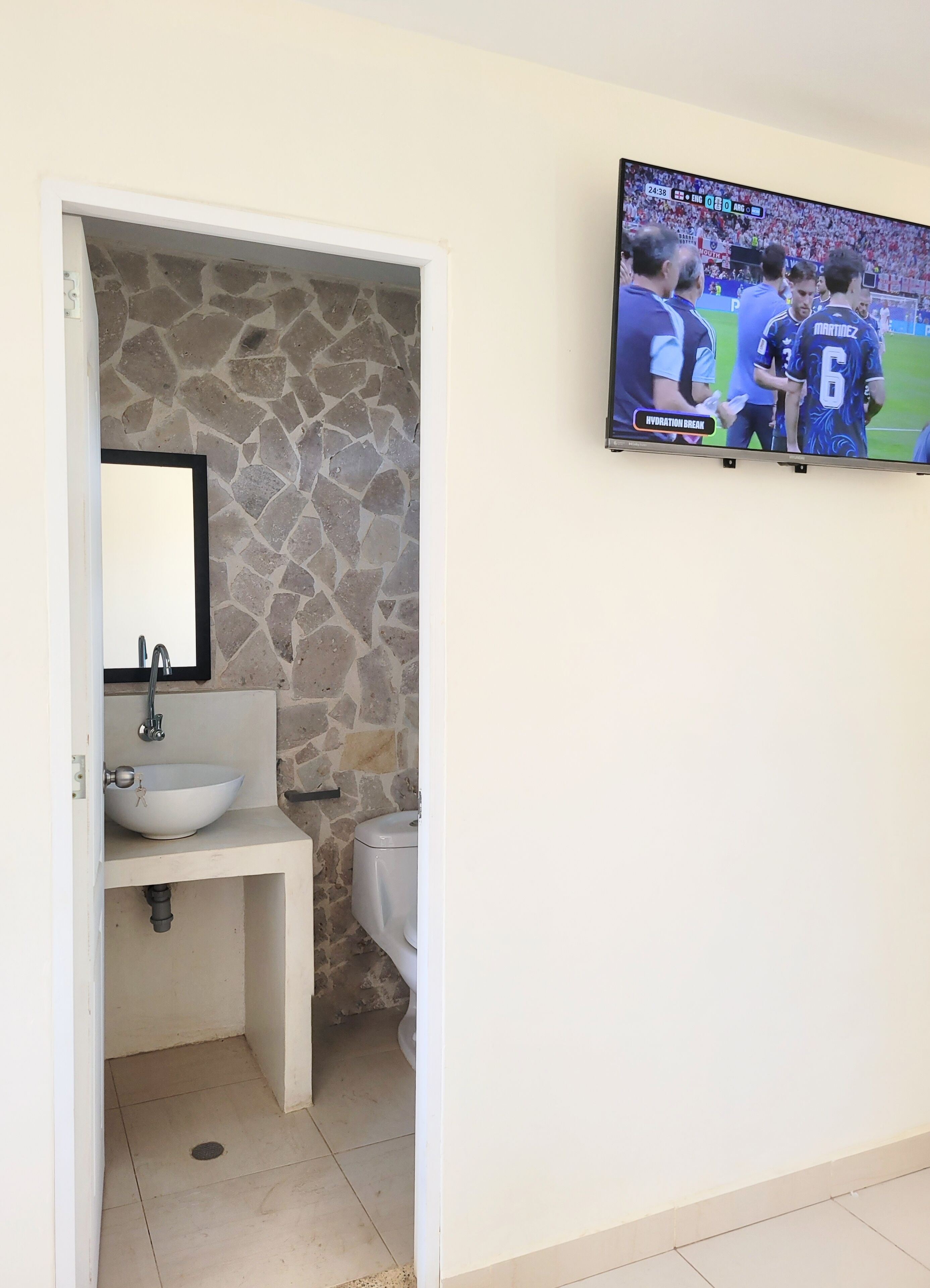 Ein Badezimmer mit Steinwand, Waschbecken, Toilette und an der Wand montiertem Fernseher, der ein Sportereignis zeigt.
