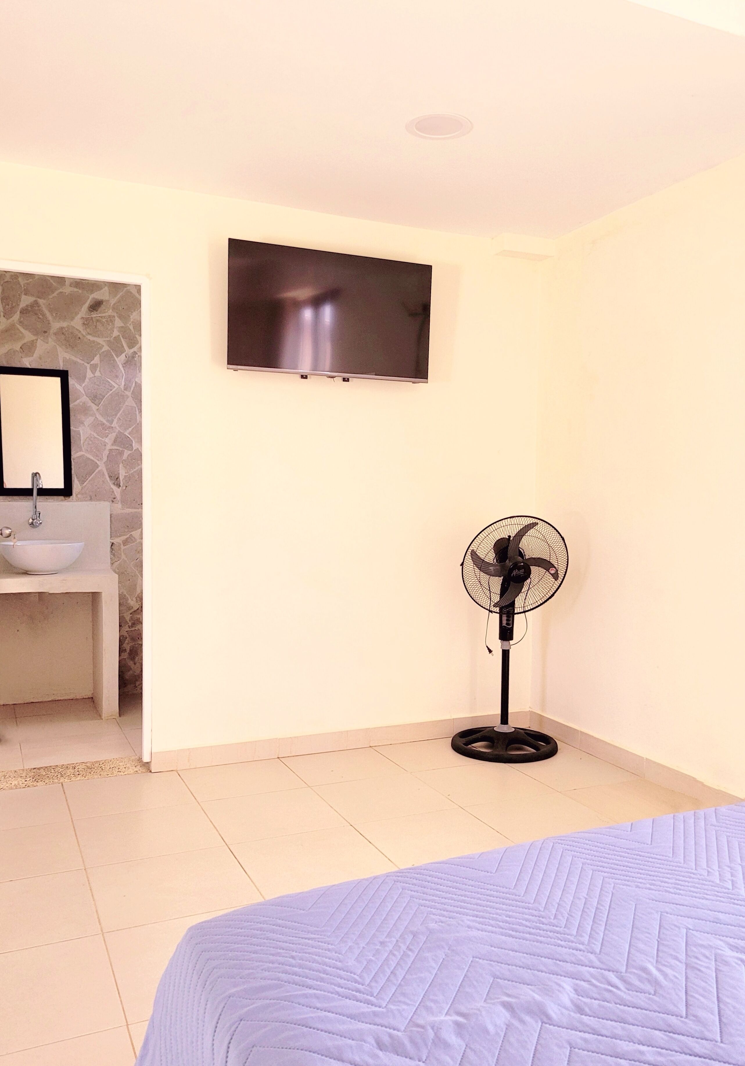 Quarto com cama, ventilador, televisão fixada na parede e banheiro com pia e espelho.