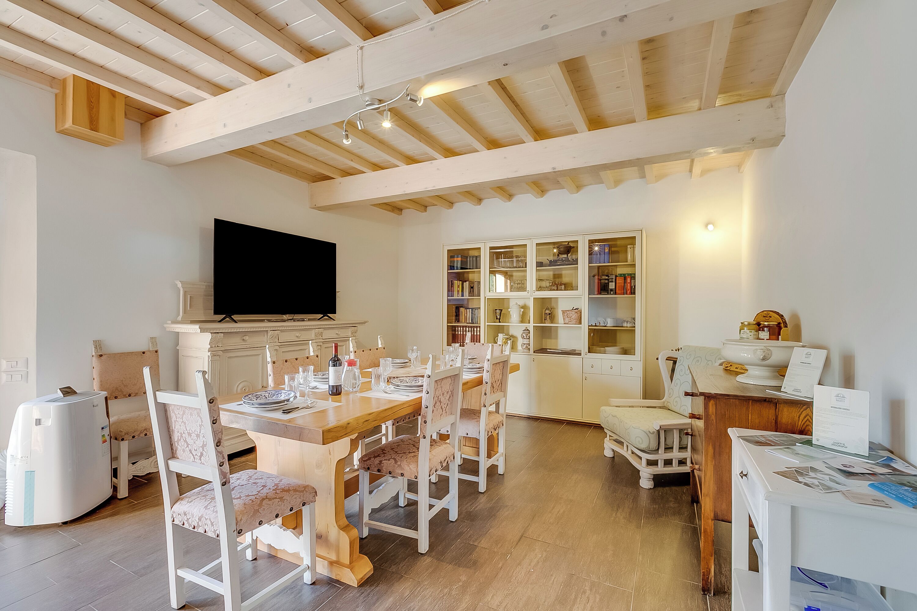 A dining area with a wooden table and chairs, a television above the table, and a kitchen area with cabinets and a refrigerator.