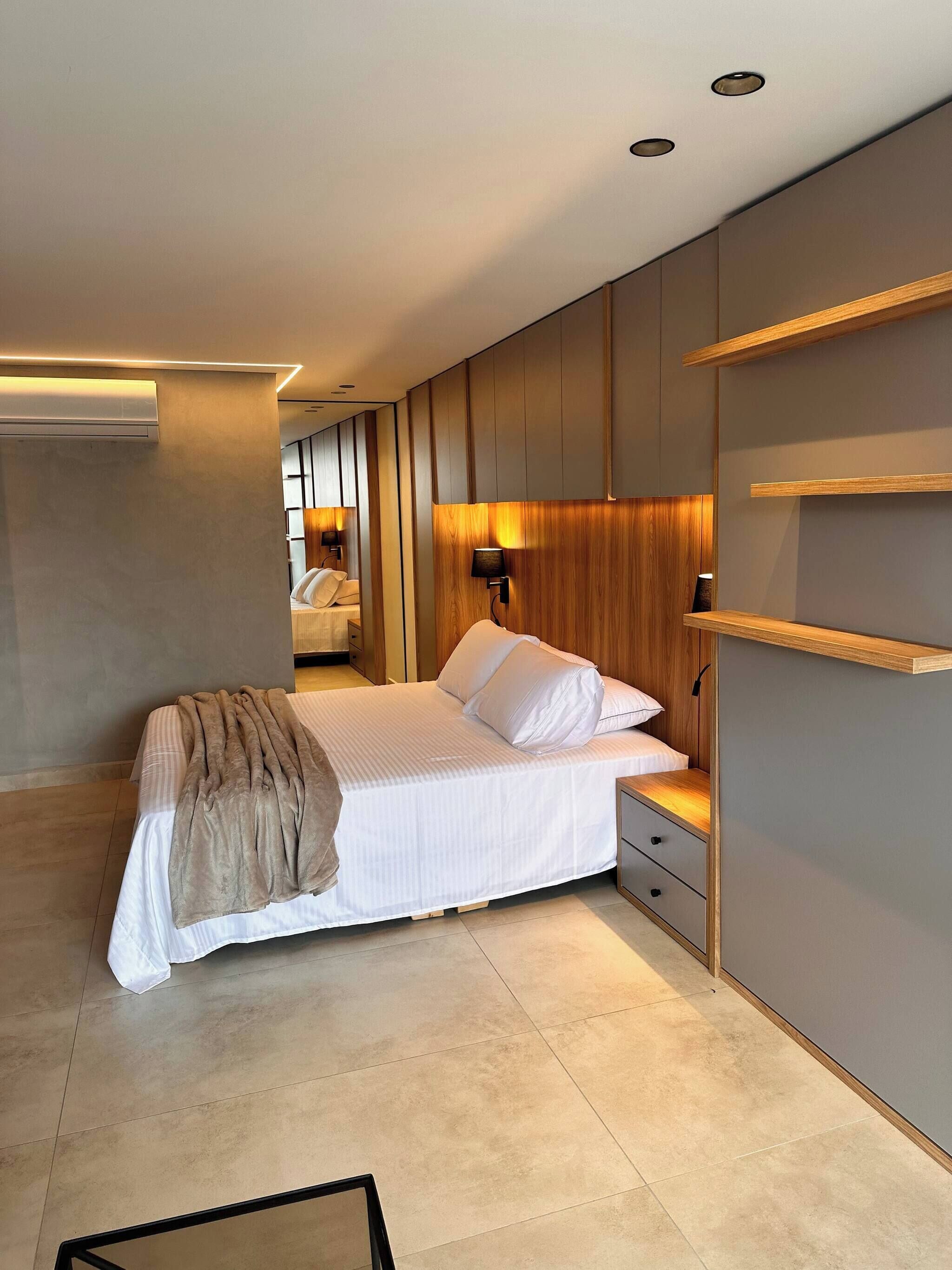 Un dormitorio moderno con una cama, mesitas de noche, una lámpara de noche, un estante en la pared, iluminación empotrada en el techo y un equipo de aire acondicionado visible.