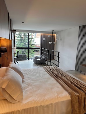 A modern bedroom with a large bed, a seating area with a chair and a small table, and a view of the outdoors through large windows. - Luxury Duplex Loft AC Rooftop Jacuzzi Laureles (Medellín)